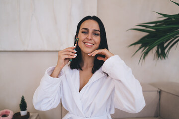Happy female smiling at camera during elegant skincare moment featuring jade roller massage...