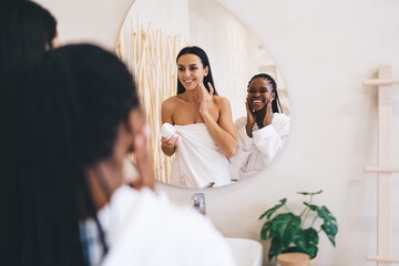 Naklejka premium Elegant bathroom scene showing personal care moment and friendship happiness. Joyful Women in white robe performing gentle facial cream massage and smiling during positive weekend spa spending