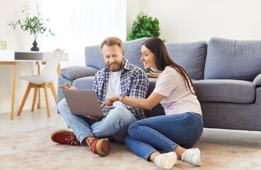 Young happy smiling couple sitting on the floor in living room at home watching news or funny video on a laptop together. Happy man and woman resting enjoying weekend. Family technology concept.