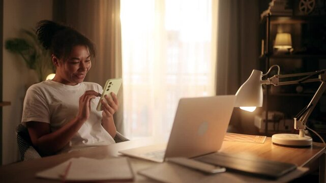 A young woman sits in a cozy home office, laughing while scrolling through content on her smartphone. She reacts joyfully, possibly watching memes or funny videos, enjoying a moment of relaxation