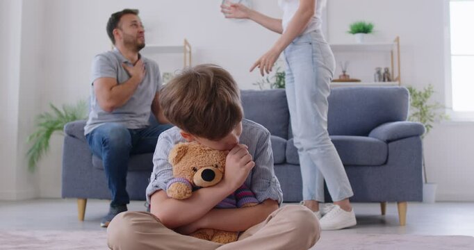 Sad child listening to fighting parents. Stressed kid boy sitting on floor in living room, holding his teddy bear while mother and father are quarreling on sofa in background. Family conflict concept 
