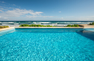 Infinite empty swimming pool with green plants next to Caribbean beach with waves and blue sky with few clouds on a sunny day in Mexico