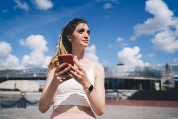 Confident young woman holding a smartphone outdoors, looking thoughtfully into the distance against...