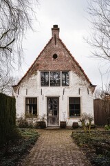 Obraz premium Charming vintage house with tall windows and a triangular roof located in a tranquil garden setting during an overcast afternoon