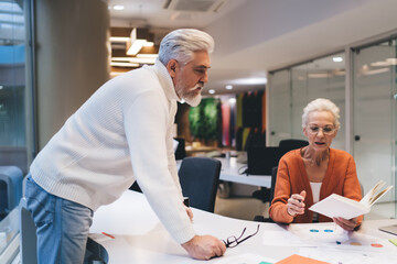 Obraz premium Senior business professionals engaged in strategic planning, man leaning over desk while woman reviews notes in a notebook. Concept of teamwork, experience, and focused collaboration in modern office