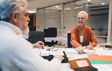 Senior woman in orange sweater smiles while discussing work with older male colleague in an office environment. Documents, charts, and notes spread across the table in a collaborative setting