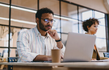 Pensive ethnic man working on laptop while sitting at table