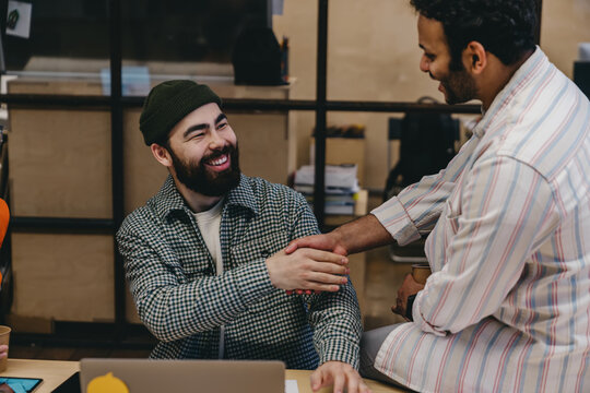 Cheerful diverse bearded male colleagues shaking hands while working in office