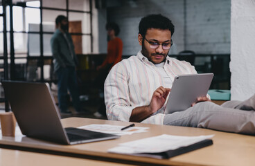 Focused ethnic man working on tablet while sitting at table with laptop