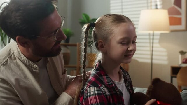 Happy Caucasian family on couch caring loving adult father does hair braid hairdo hairstyle coiffure hair cute daughter dad man talking with kid child playing toy Teddy bear at home bonding adopt