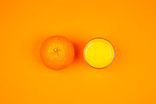 Een sinaasappel en een glas sinaasappelsap op een oranje achtergrond. De felle kleuren en minimalistische compositie benadrukken de frisheid, versheid en gezonde uitstraling van het fruit en sap.