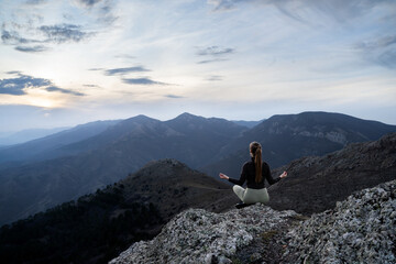 Fototapeta premium Unrecognizable traveler woman with a ponytail sitting in a lotus position, meditation in the mountains. Mindfulness, connection with nature, inner peace, spiritual balance. Blue hour. Rear view.