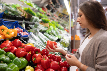 Woman selecting fresh red bell peppers in the vegetable section of a supermarket. Concept of healthy eating, buying natural products, and caring about food quality.