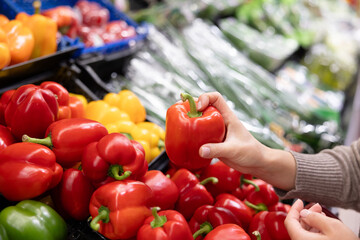 A woman's hand selects fresh red bell peppers in the vegetable section of a supermarket. The concept of choosing quality ingredients for cooking