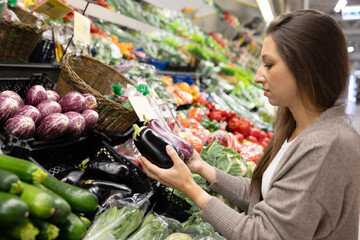 Woman examining a striped eggplant in the produce section of a supermarket. Concept of selecting fresh vegetables, making healthy food choices, and mindful grocery shopping