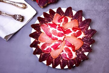 Sliced meat on a plate, knife and tongs on a white napkin, on a gray background