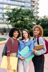 Vertical shot of three young university female students smiling together at camera on campus, holding folders and backpacks, enjoying their university experience. Education lifestyle and youth concept