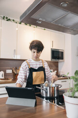 Teenage girl stirring a pot and reading recipe on a digital tablet while cooking in a modern kitchen