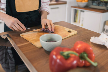 Unrecognizable teenage girl in apron slicing olives on wooden cutting board, preparing healthy meal with fresh vegetables in modern kitchen