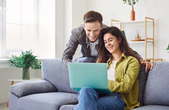 Young family couple sitting at home, sofa looking smiling at laptop screen, happy young married spouse web surfing, making purchases online booking flight tickets, reading good news, leisure time