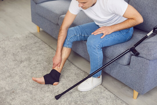 Young man sitting on the sofa with a crutch and massaging his leg, foot and sprained ankle wrapped in a medical joint stabilizer brace bandage with adjustable straps. Injury and pain treatment concept