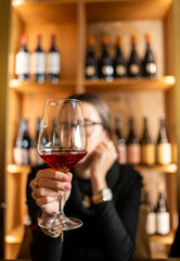 A young woman holds a fine crystal glass of wine in the foreground with a wine cellar from Tuscany on a large unfocused wood shelves. Concept of tasting, wine library, delicacy and sophistication
