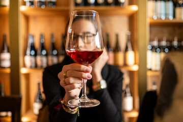 A young woman holds a fine crystal glass of wine in the foreground with a wine cellar from Tuscany on a large unfocused wood shelves. Concept of tasting, wine library, delicacy and sophistication