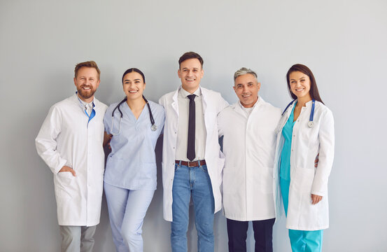 Group of young and adult doctors clinicians and cardiologists in scrubs and white coats standing together, hugging each other and smiling at camera on gray background. Happy clinic or hospital staff.