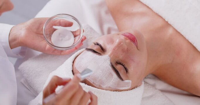 Beautician applying anti-aging facial cream on woman client face to prevent wrinkles in spa salon. Skin care, cosmetic procedures for facial care and beauty treatment concept. 4k video.