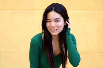 Portrait of a smiling young Japanese woman touching her hair, wearing a green shirt on a yellow...