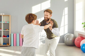 Obraz premium Happy smiling young man with child boy wearing sportswear doing boxing sport exercise in gym during workout. Father and son exercising together. Family health and body care concept.