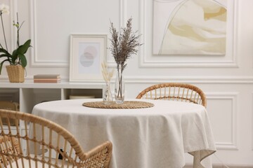 Table with white tablecloth, dried flowers in vases and chairs in stylish room. Interior design