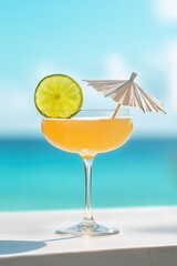 Orange cocktail with lime slice and cocktail umbrella standing on white table with blurred turquoise sea in background