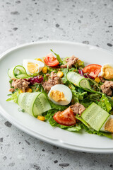 Fresh salad with tuna, boiled eggs, cucumbers, tomatoes, and corn on a white plate, beautifully arranged on a speckled gray surface, ideal for healthy eating concepts