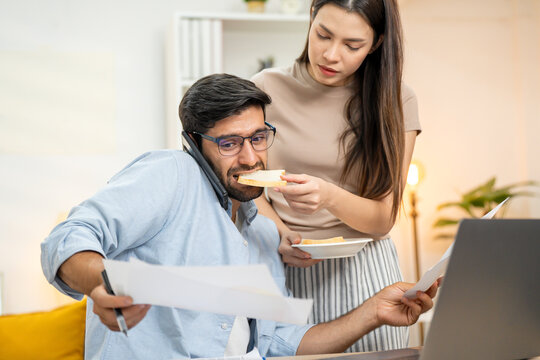 Young businessman working at home office on laptop computer, finishing his online work, while his girlfriend and roommate sitting behind on sofa relaxing and waiting. Freelance business. couple.