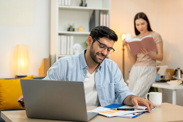 Fototapeta premium Young businessman working at home office on laptop computer, finishing his online work, while his girlfriend and roommate sitting behind on sofa relaxing and waiting. Freelance business. couple.