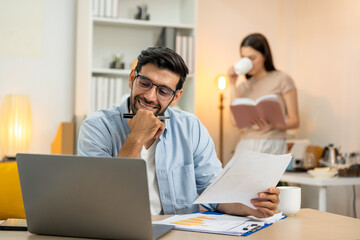 Obraz premium Young businessman working at home office on laptop computer, finishing his online work, while his girlfriend and roommate sitting behind on sofa relaxing and waiting. Freelance business. couple.