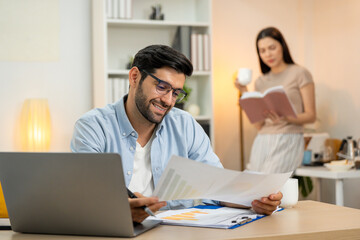 Young businessman working at home office on laptop computer, finishing his online work, while his girlfriend and roommate sitting behind on sofa relaxing and waiting. Freelance business. couple.
