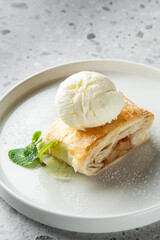 A delicious slice of pastry dessert with layers, topped with a scoop of vanilla ice cream and garnished with a fresh mint leaf, presented on a white plate.