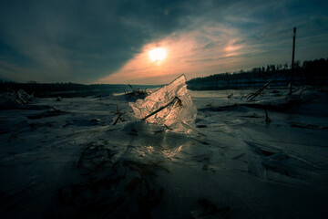 A toned photograph captures a piece of ice standing on the frozen surface of the river, with sunlight passing through it. Some parts of the river remain ice-free.