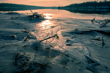 A toned picture a piece of ice standing on the frozen surface of the river, with evening sunlight passing through it. Some parts of the river remain ice-free.