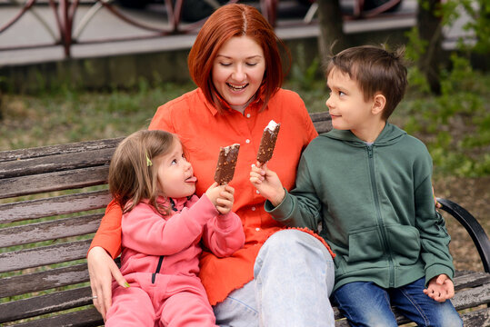 A happy mother with son and daughter sharing ice cream sitting on bench on a street. Little girl playfully sticks out her tongue, teasing mom with ice-cream bar. Family having a good time together. 