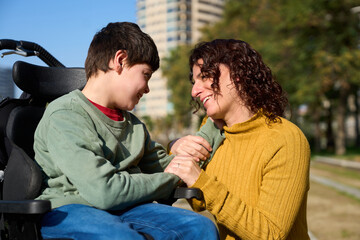 Happy mother holding hands with her son with disability in wheelchair outdoors in a park - cerebral...