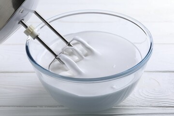 Mixing whipped cream with hand mixer in bowl on white wooden table, closeup