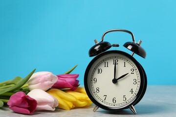 Spring time. Alarm clock and beautiful tulips on grey marble table against light blue background