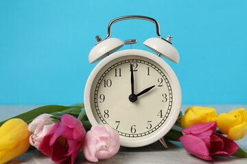 Spring time. Alarm clock and beautiful tulips on grey marble table against light blue background,...