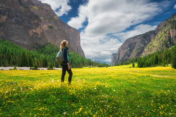 Girl with backpack on the meadow with yellow flowers and green grass in beautiful alpine mountain valley at sunset in summer.Young woman in alps, sky with clouds. Hiking. Spring in Dolomites, Italy