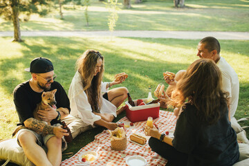Happy people having picnic with hot dogs and cold drinks, sitting on a plaid in a park.