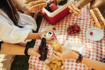 Happy people having picnic with hot dogs and cold drinks, sitting on a plaid in a park.