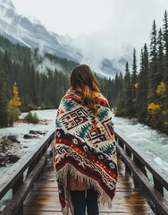A woman wrapped in a pure white blanket with colorful black and red Native American patterns stands on a wooden bridge over a river against a backdrop of high mountains.